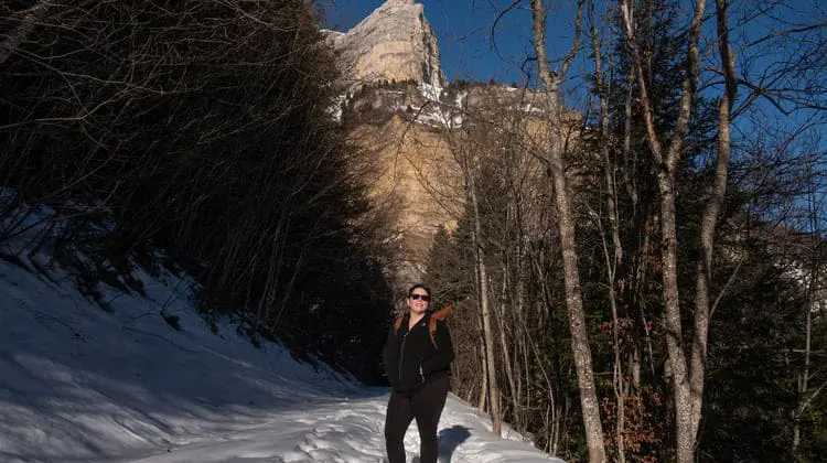 Mulher de óculos escuro em trilha nevada em frente aos Alpes.