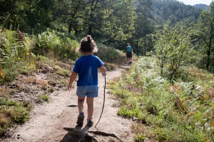Criança pequena em primeiro plano caminhando com um graveto. Ao fundo pai. A cena é em uma trilha com vegetação verde.