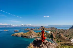 Mulher em uma pedra, num dia de sol, com o mar no fundo, enquanto segura o seu chapéu