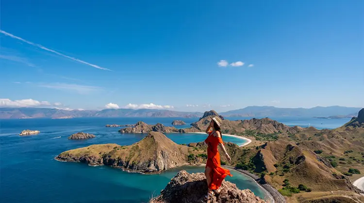 Mulher em uma pedra, num dia de sol, com o mar no fundo, enquanto segura o seu chapéu