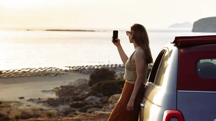 Mulher com o celular na mão, encostada em um carro, tirando foto da paisagem