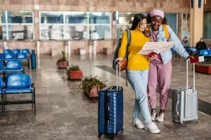 Duas jovens turistas sorridentes olhando para um mapa, segurando malas de rodinha, dentro de um saguão.