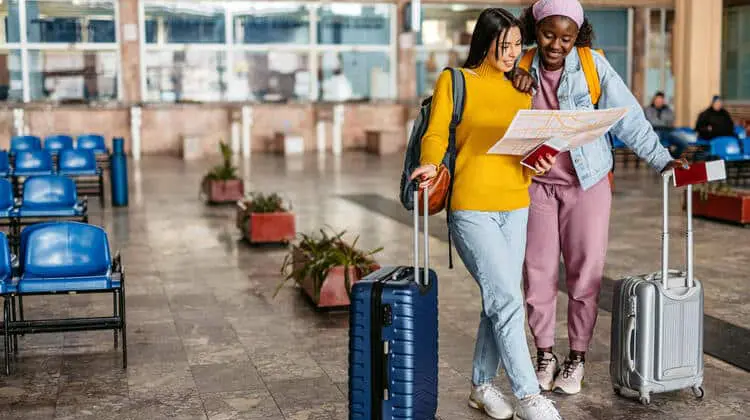 Duas jovens turistas sorridentes olhando para um mapa, segurando malas de rodinha, dentro de um saguão.