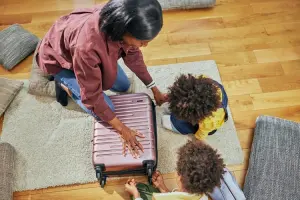 Mãe abaixada no chão com filhos, arrumando a mala de viagem das crianças.