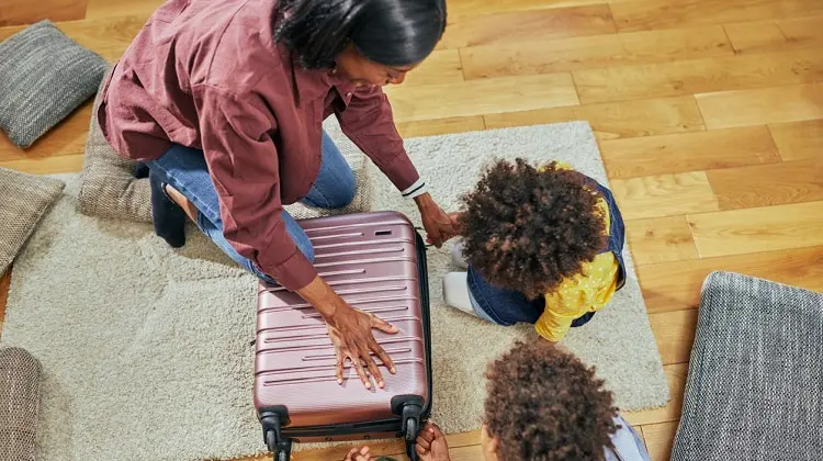 Mãe abaixada no chão com filhos, arrumando a mala de viagem das crianças.