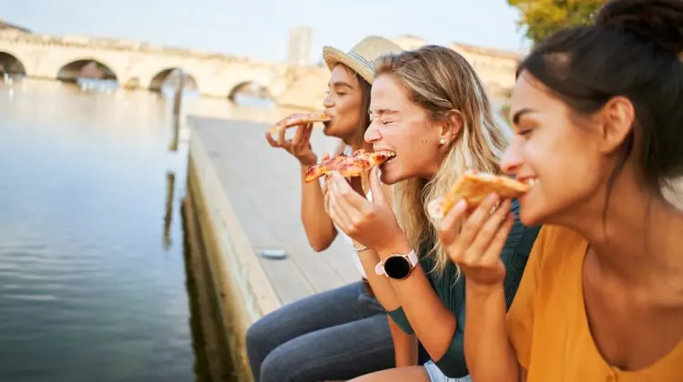 Três mulheres comendo pizza na beira de um rio