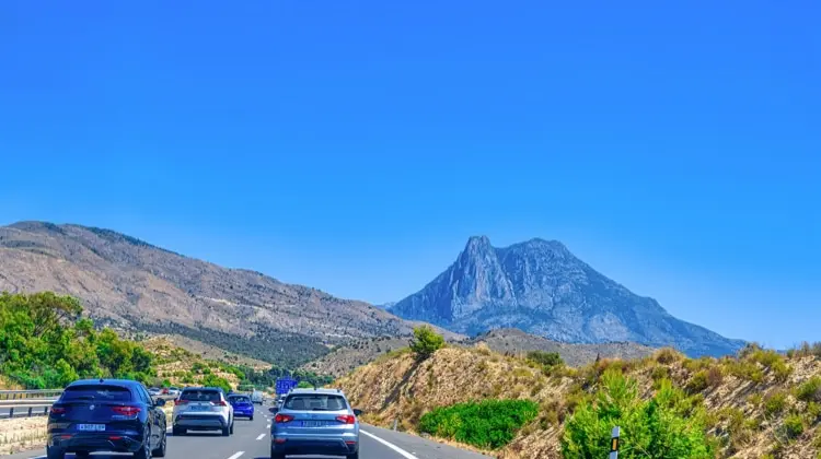 Estrada espanhola com carros passando e uma montanha ao fundo