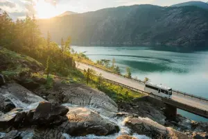 Ônibus branco em estrada vista de cima, paisagem com lago e montanha, sol ao fundo, paisagem com pouca neve, na Noruega
