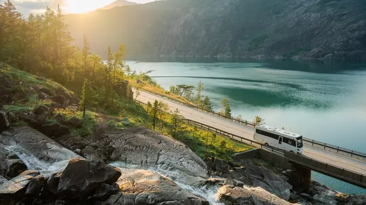 Ônibus branco em estrada vista de cima, paisagem com lago e montanha, sol ao fundo, paisagem com pouca neve, na Noruega