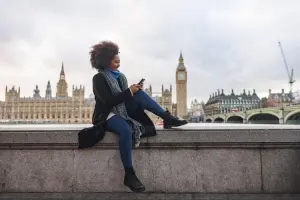Mulher mexe no celular em dia nublado e ao fundo o icônico Big Ben, parte do Palácio de Westminster, aparece imponente na paisagem urbana de Londres.
