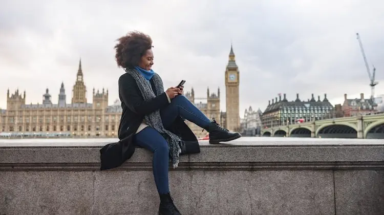 Mulher mexe no celular em dia nublado e ao fundo o icônico Big Ben, parte do Palácio de Westminster, aparece imponente na paisagem urbana de Londres.