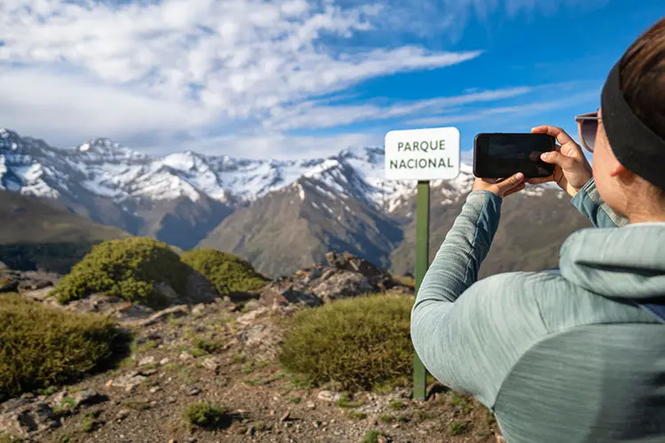 Pessoa fotografando placa de parque nacional com montanhas ao fundo.