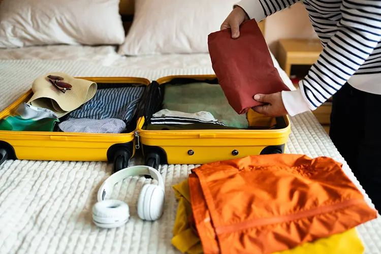 Pessoa organizando mala em quarto de hotel, com roupas e acessórios visíveis ao lado da mala.