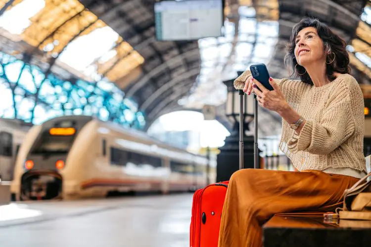 Mulher sentada em estação de trem com mala vermelha e celular na mão