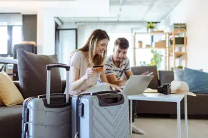 Casal abre com conta internacional para viagem no laptop, ao lado de malas prontas.