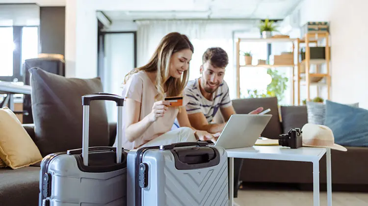 Casal abre com conta internacional para viagem no laptop, ao lado de malas prontas.