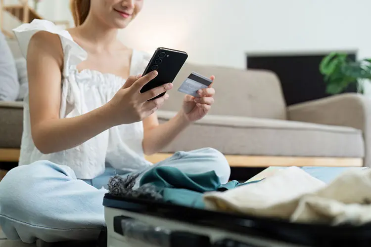 Mulher acessa conta no celular enquanto arruma a mala.
