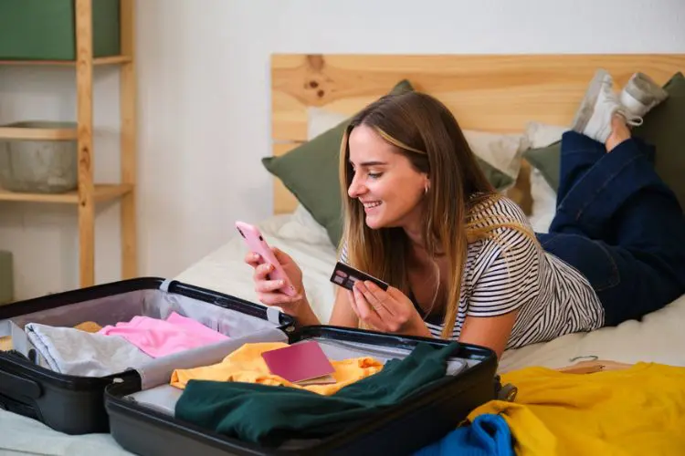 Mulher deitada na cama usa o celular com um cartão na mão e uma mala aberta à sua frente