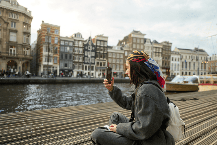 Mulher sentada à beira de um canal em cidade europeia, usando celular para fazer fotos.