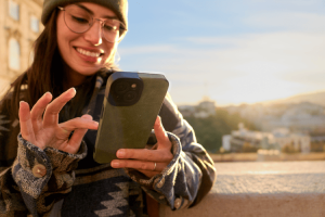 Mulher usando smartphone ao ar livre durante o pôr do sol, vestindo óculos, gorro e casaco estampado.
