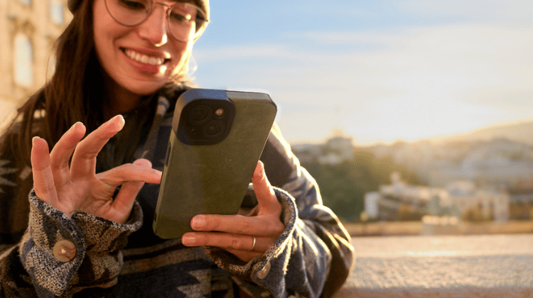 Mulher usando smartphone ao ar livre durante o pôr do sol, vestindo óculos, gorro e casaco estampado.