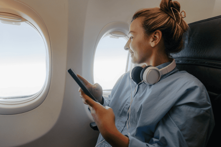 Mulher sentada em poltrona de avião olhando pela janela com um celular na mão e fones de ouvido no pescoço.