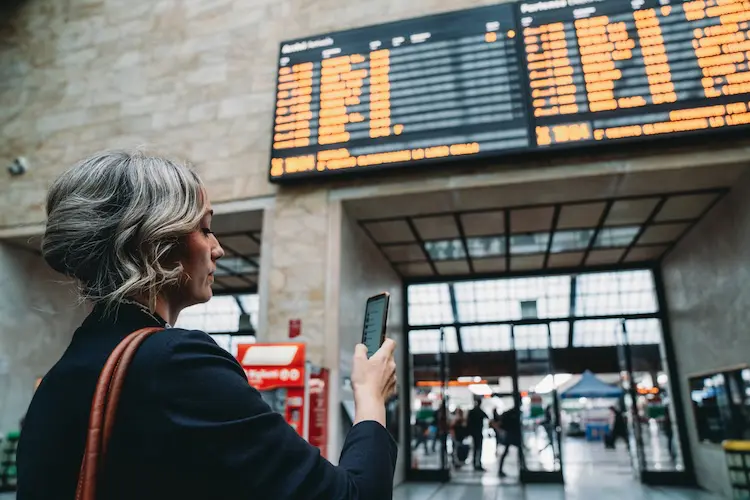 Mulher olhando para o celular enquanto observa o painel eletrônico de horários em estação de transporte.