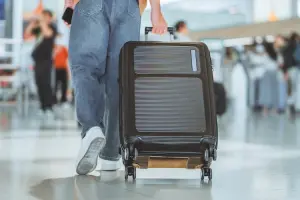 Pessoa puxando uma mala Luxcel preta de rodinhas em aeroporto movimentado.