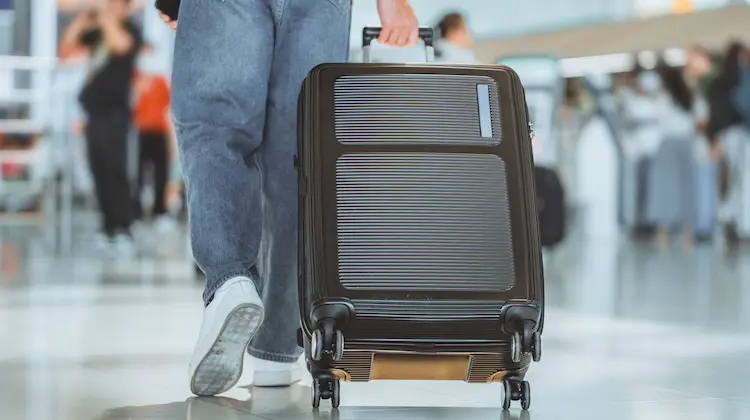 Pessoa puxando uma mala Luxcel preta de rodinhas em aeroporto movimentado.