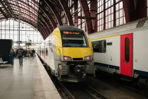 Trem de passageiros amarelo e branco parado em estação coberta, com pessoas caminhando na plataforma.