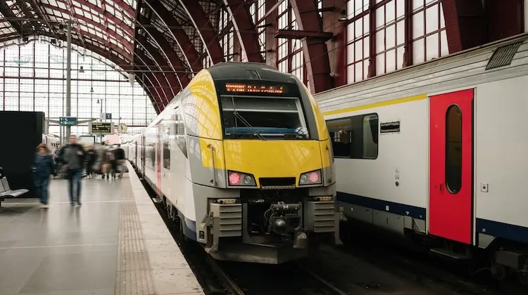 Trem de passageiros amarelo e branco parado em estação coberta, com pessoas caminhando na plataforma.