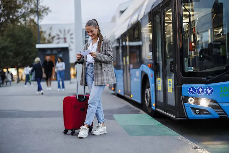 Mulher sorrindo enquanto segura passagens, ao lado de mala vermelha e ônibus azul estacionado.