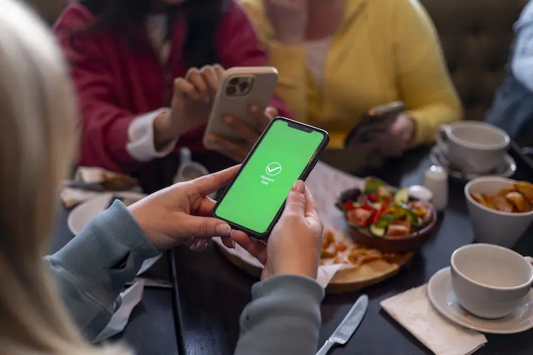 Pessoa segura celular com tela verde exibindo confirmação de pagamento, enquanto está à mesa com outras pessoas usando smartphones.