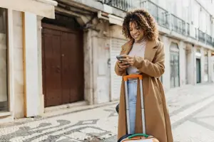 Mulher viajante sorridente olha o celular enquanto segura uma mala de rodinhas em uma rua de calçada portuguesa.
