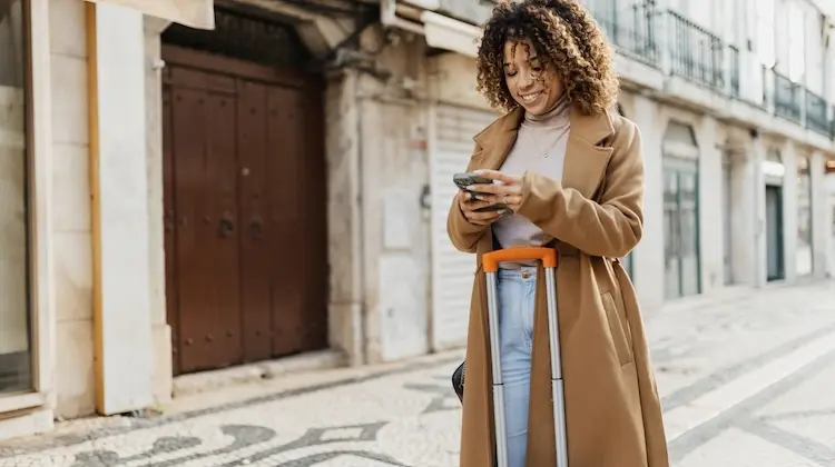 Mulher viajante sorridente olha o celular enquanto segura uma mala de rodinhas em uma rua de calçada portuguesa.