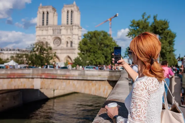 Mulher fotografa a Catedral de Notre-Dame, em Paris, segurando um café e o celular às margens do rio Sena.