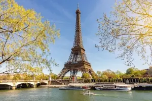 Torre Eiffel em Paris em um dia ensolarado, vista da margem do rio Sena com barcos passando.