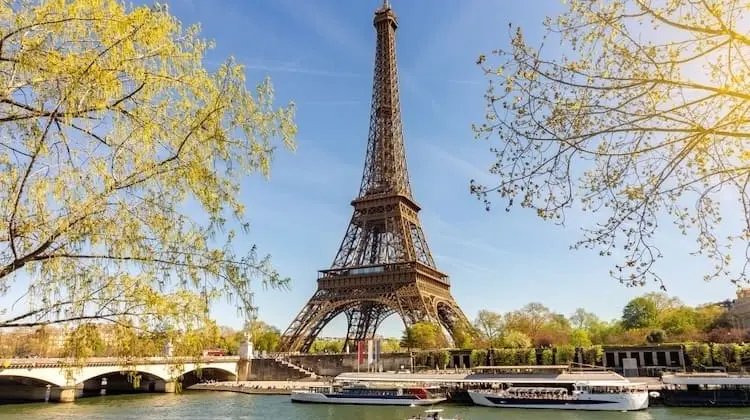 Torre Eiffel em Paris em um dia ensolarado, vista da margem do rio Sena com barcos passando.