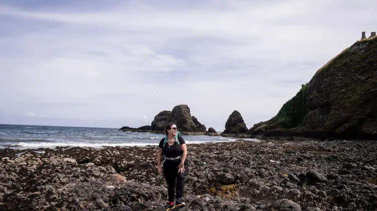 Mulher com mochila em frente a rochas e mar da Escócia.