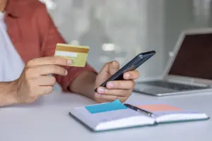 Pessoa segurando um cartão e um celular enquanto faz uma transação online, com notebook e caderno sobre a mesa.