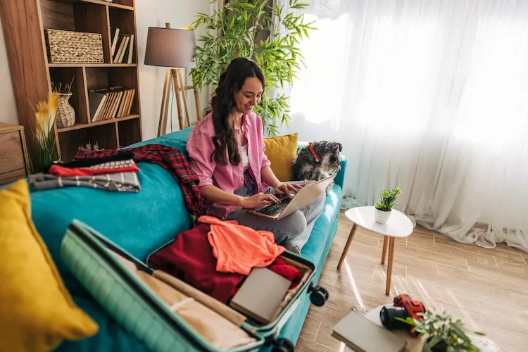 Mulher sentada no sofá com uma mala aberta e um notebook no colo enquanto planeja uma viagem.