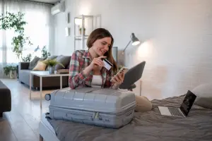 Mulher sentada na cama usando o celular e segurando um cartão, com uma mala de viagem à frente.