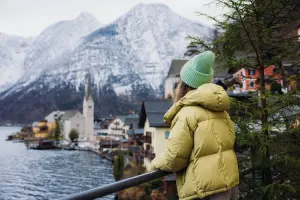 Pessoa usando casaco de inverno observa vila europeia à beira de um lago com montanhas nevadas ao fundo.