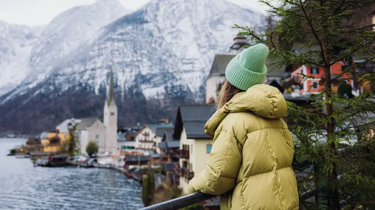 Pessoa usando casaco de inverno observa vila europeia à beira de um lago com montanhas nevadas ao fundo.