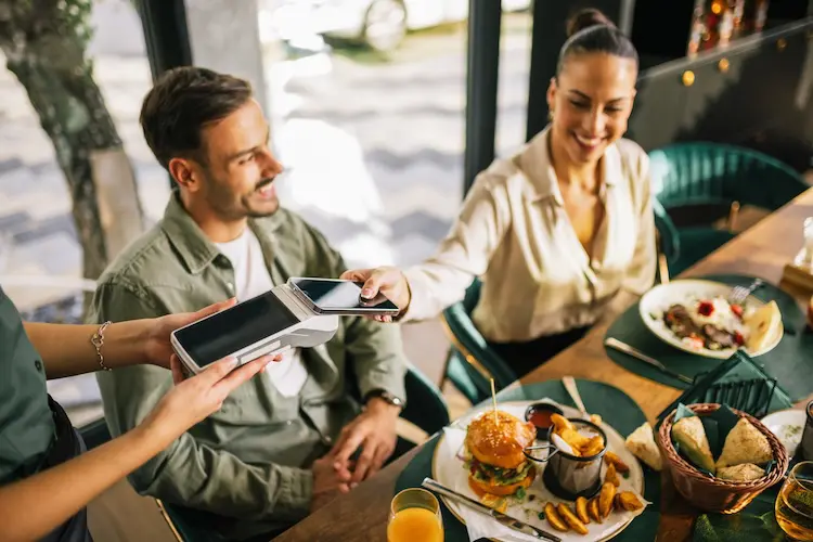 Mulher pagando a conta em um restaurante usando o celular por aproximação, enquanto um homem sorri ao lado.