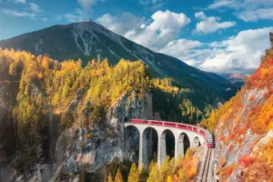 Trem vermelho Bernina Express em contraste com as árvores douradas passando em viaduto em curva.
